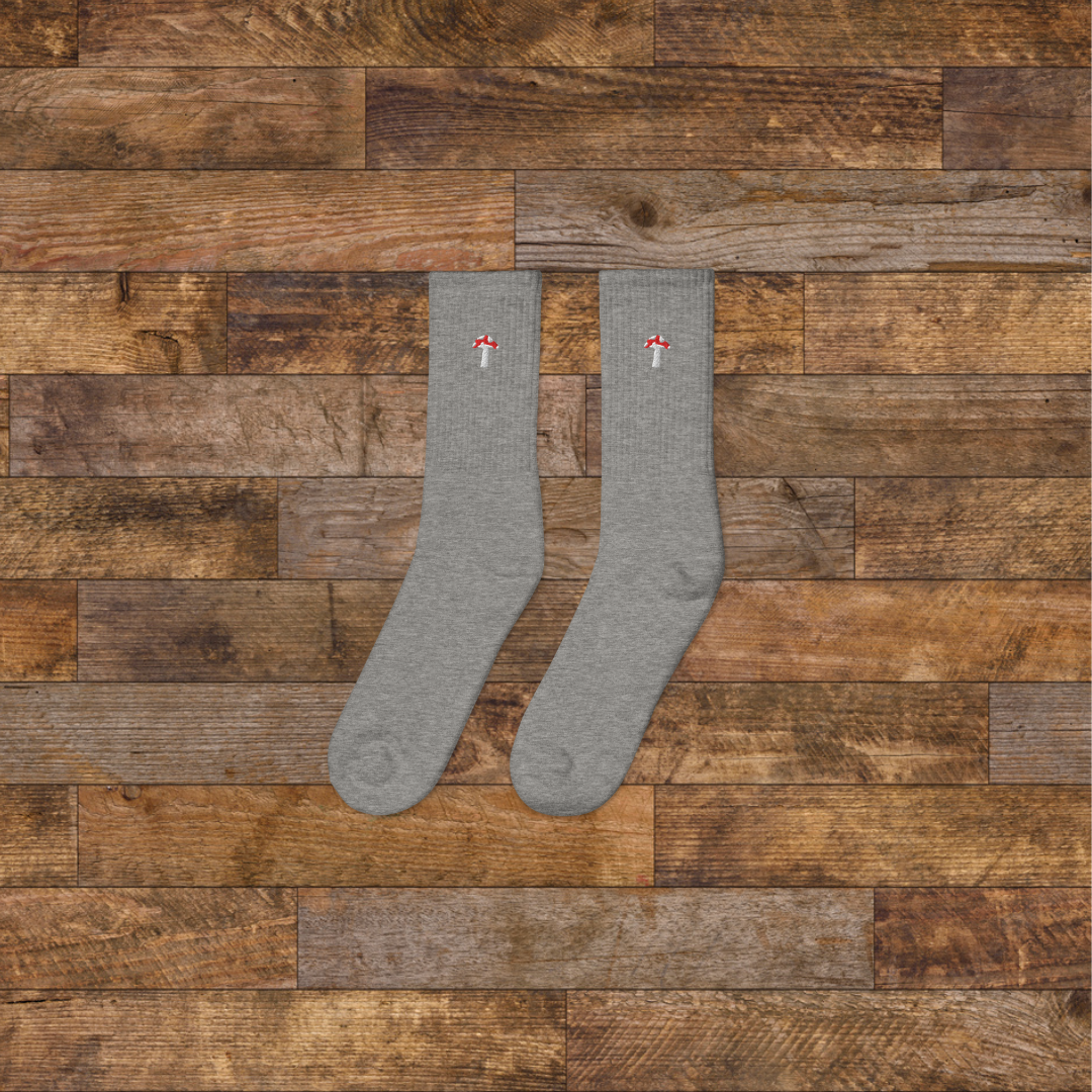 left grey Embroidered Mushroom Crew Socks - ribbed knit with fly agaric red mushroom ankle design, wooden background.