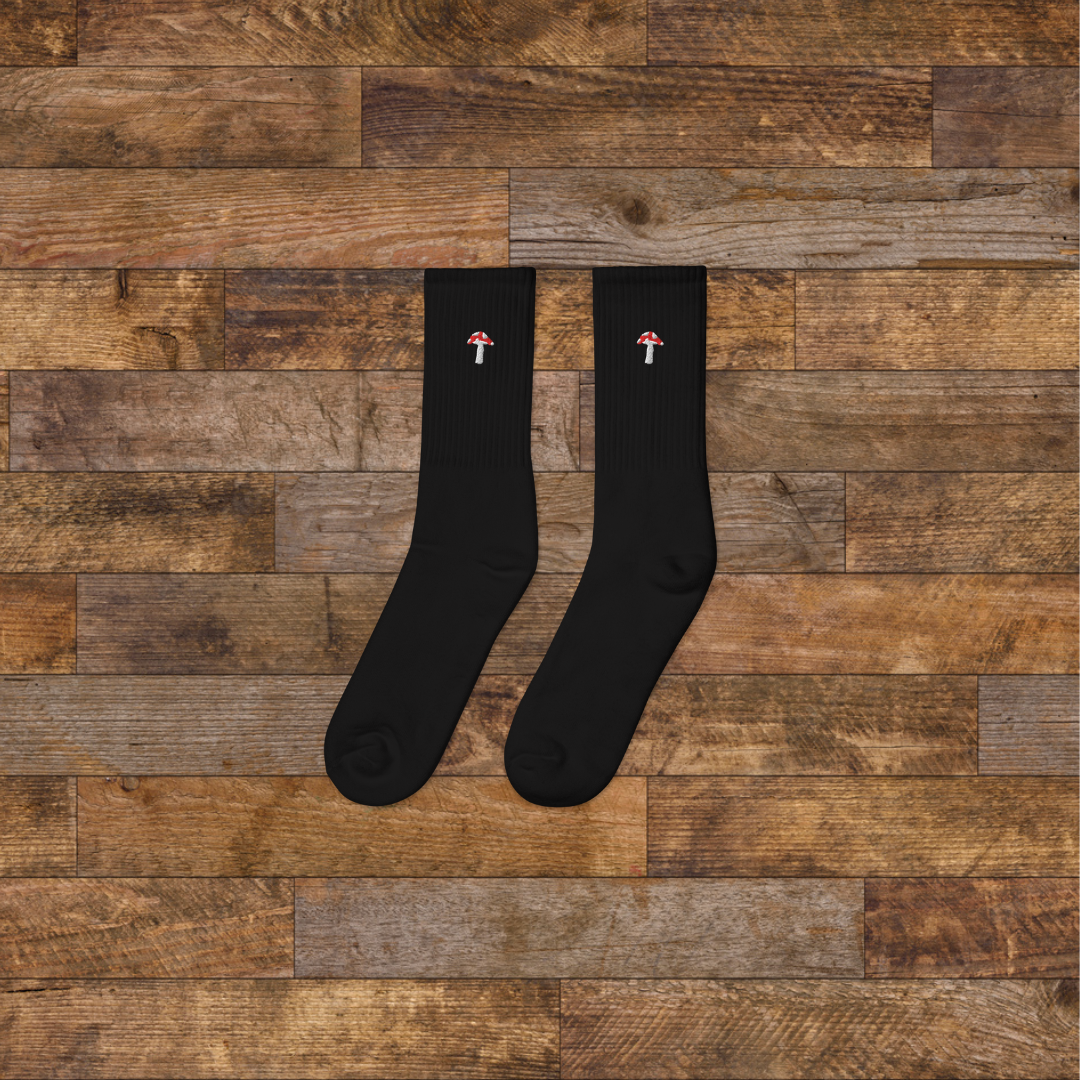 left black Embroidered Mushroom Crew Socks - ribbed knit with fly agaric red mushroom ankle design, wooden background.