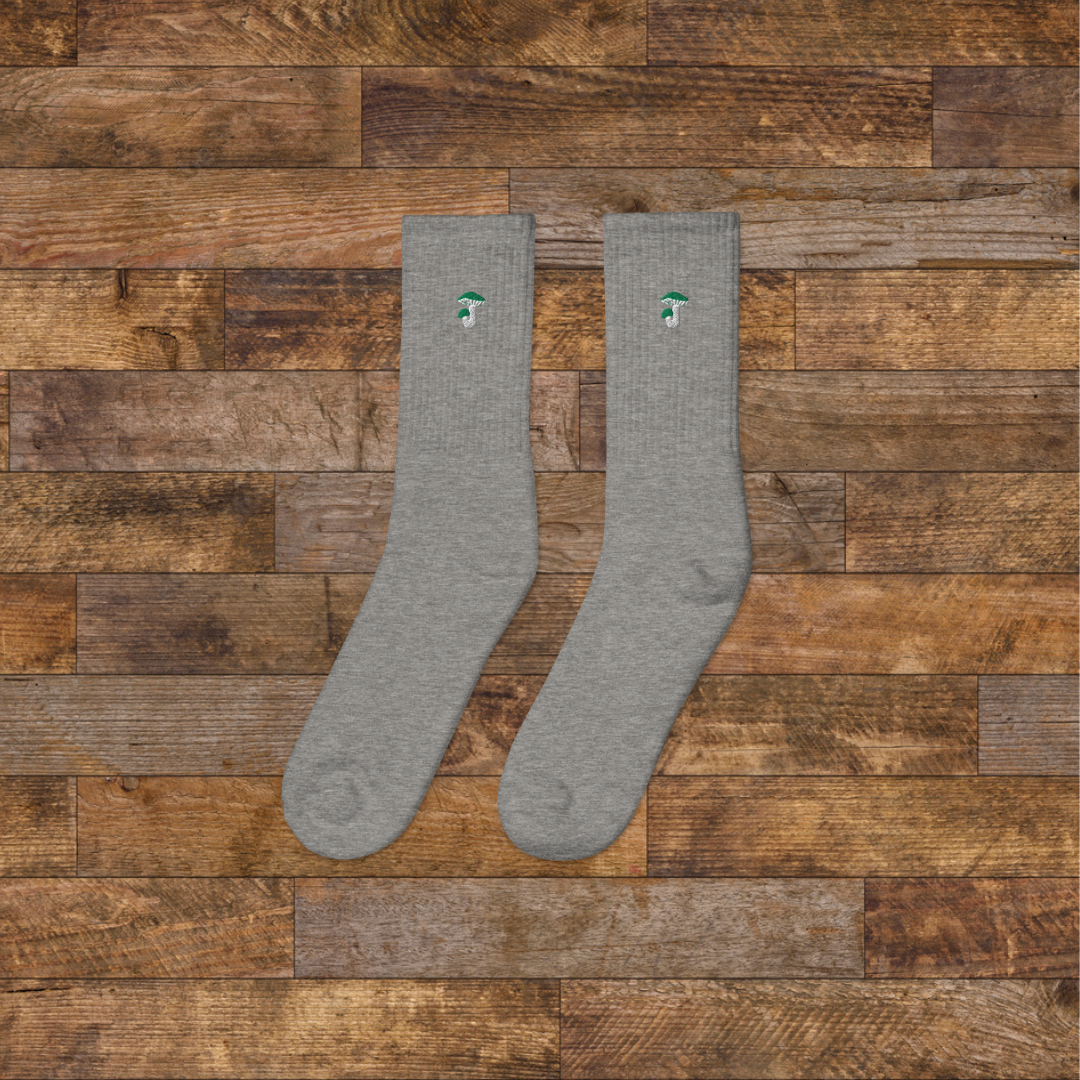 Left grey Embroidered Mushroom Crew Socks - ribbed knit with Russula green mushroom ankle design, wooden background.