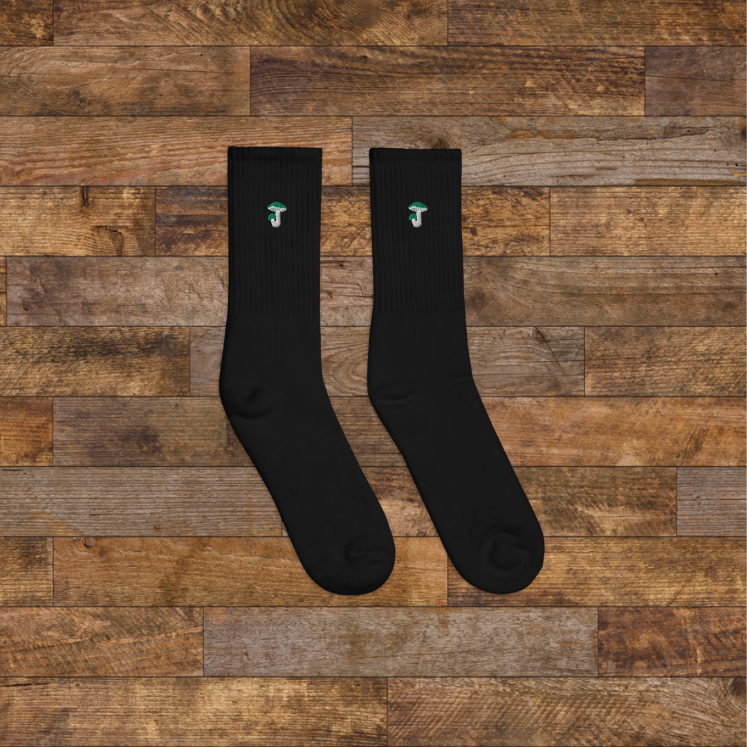 Right black Embroidered Mushroom Crew Socks - ribbed knit with Russula green mushroom ankle design, wooden background.