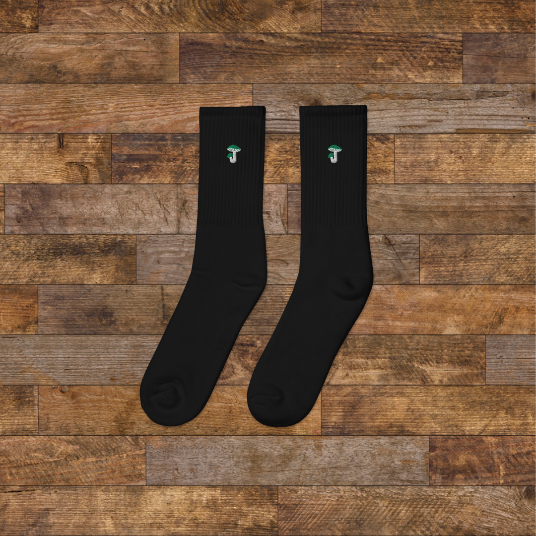 Left black Embroidered Mushroom Crew Socks - ribbed knit with Russula green mushroom ankle design, wooden background.