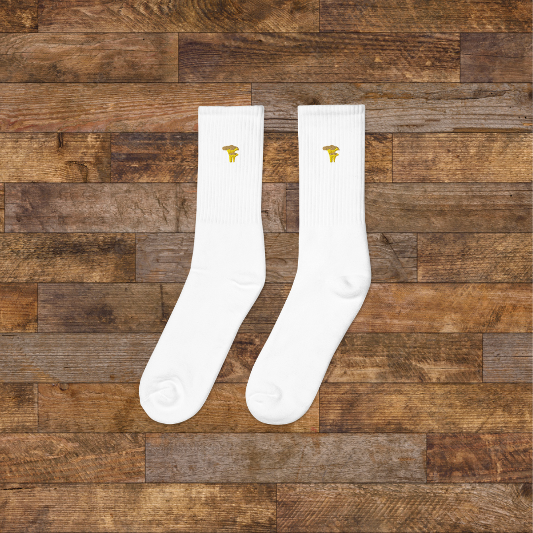 Left white Embroidered Mushroom Crew Socks - ribbed knit with Chanterelle yellow mushroom ankle design, wooden background.