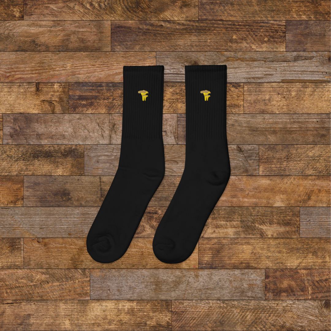 Left Black Embroidered Mushroom Crew Socks - ribbed knit with Chanterelle yellow mushroom ankle design, wooden background.