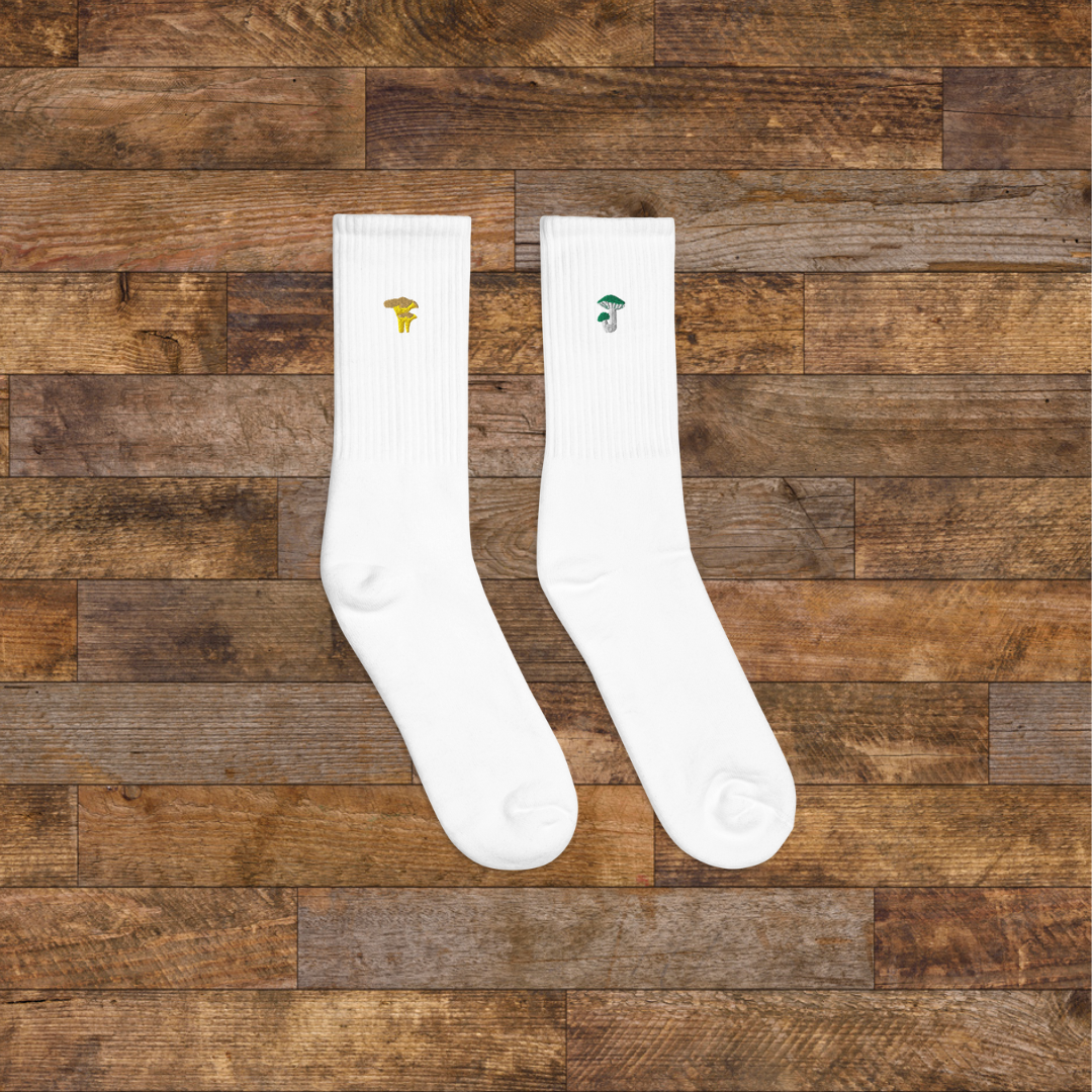 right white Embroidered Mushroom Crew Socks - ribbed knit with russula green and chanterelle yellow mushroom ankle design, wooden background.