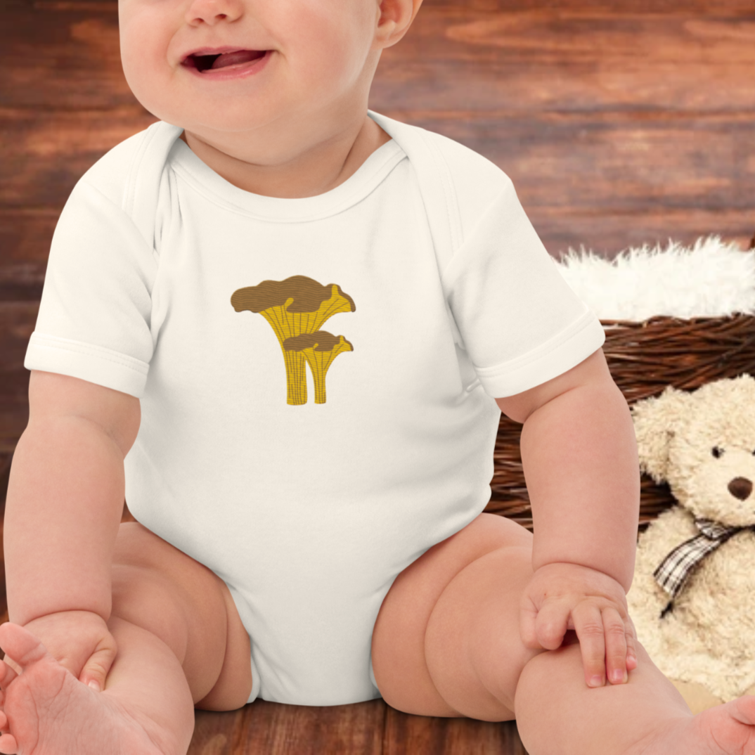 Baby wearing a white baby body with a chanterelle mushroom. Teddy bear in the background