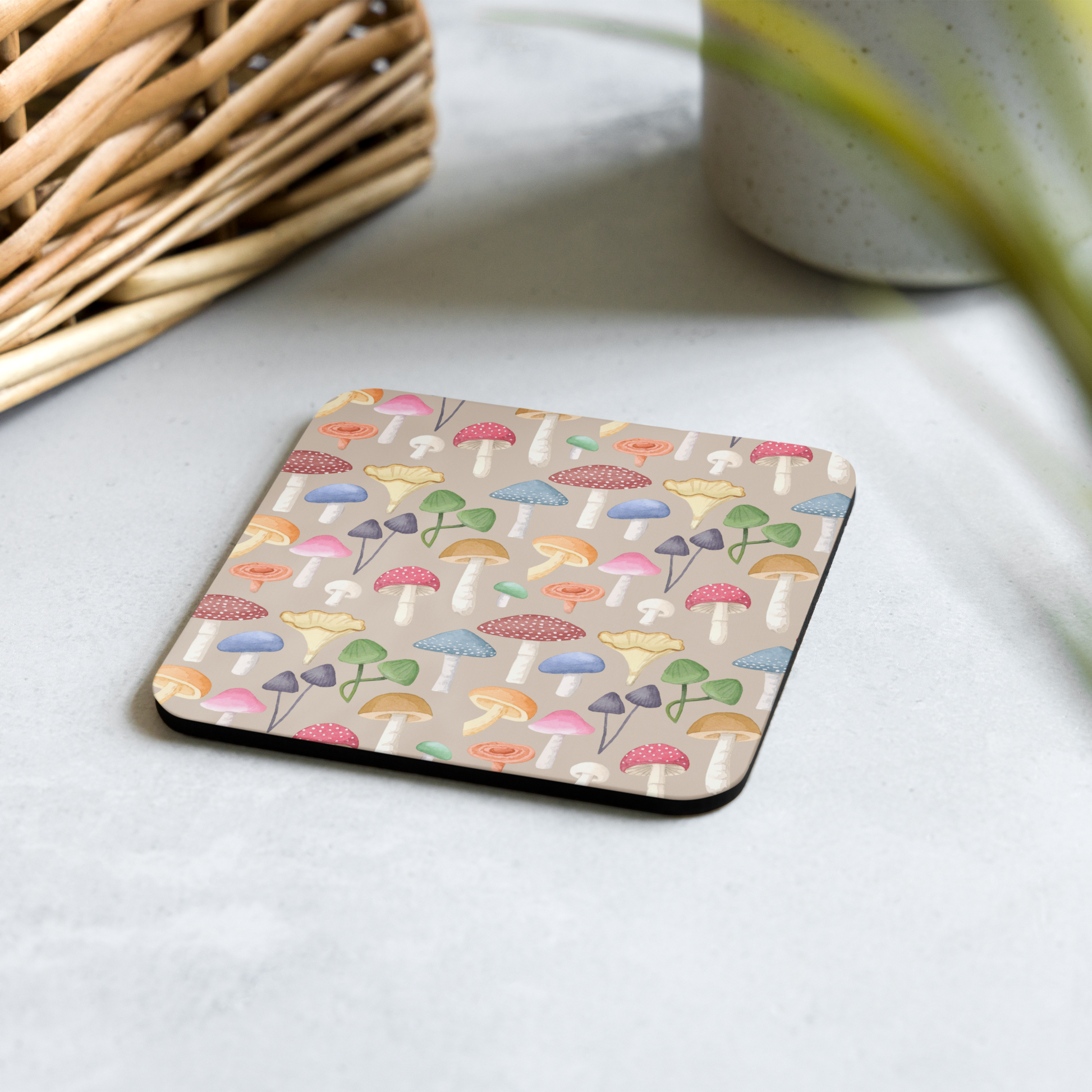 A square beige cork-back coaster with a multicolored mushroom pattern, placed on a table.