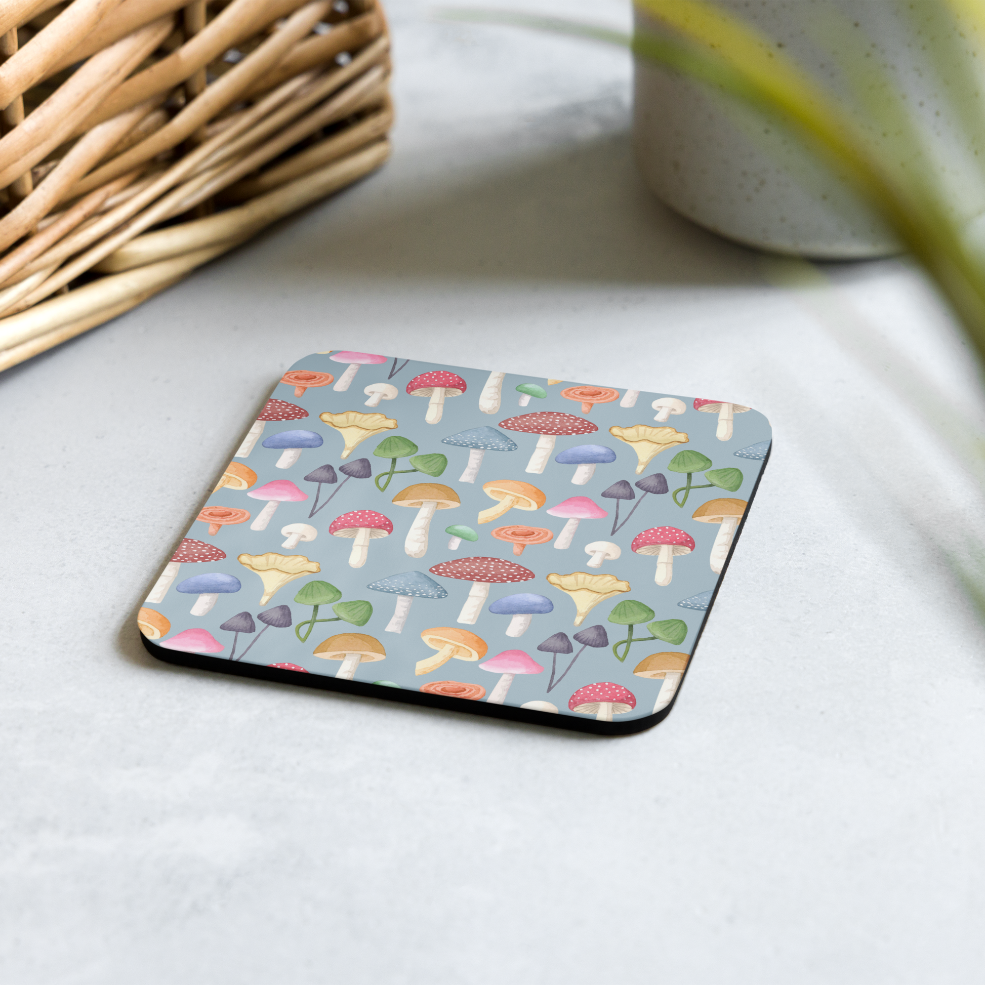 A square coaster with a mushroom pattern placed on a table surface, with a basket and a plant in the background.