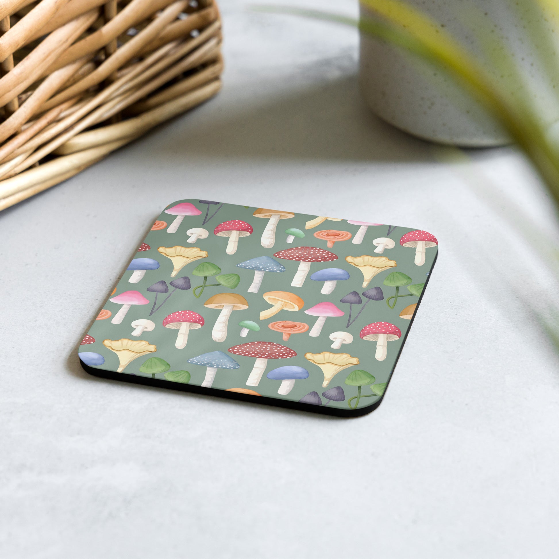 A square green cork-back coaster with a pattern of colorful mushrooms displayed on a table.