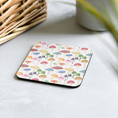 A square white cork-back coaster with a multicolored umbrella pattern, placed on a table surface next to a basket and a plant.