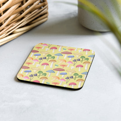 A square yellow cork-back coaster with a multicolored mushroom pattern, placed on a table.